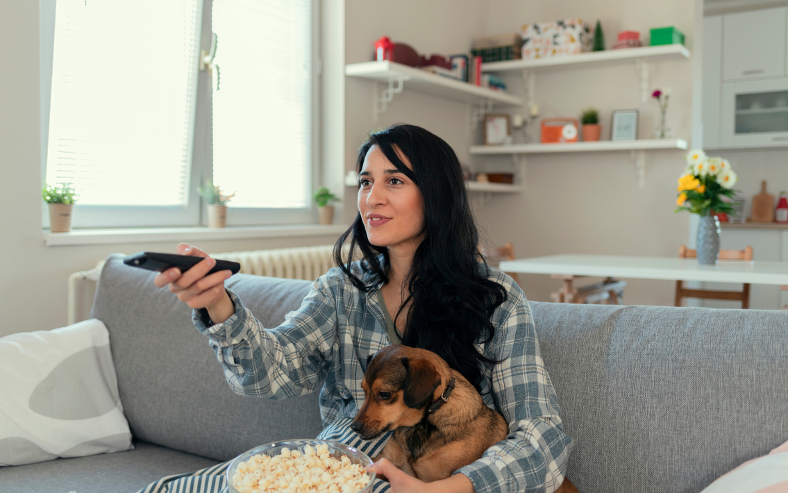 An image of a long, black haired woman wearing a green plaid button down pointing a remote while holding popcorn and a tan chihuahua. 